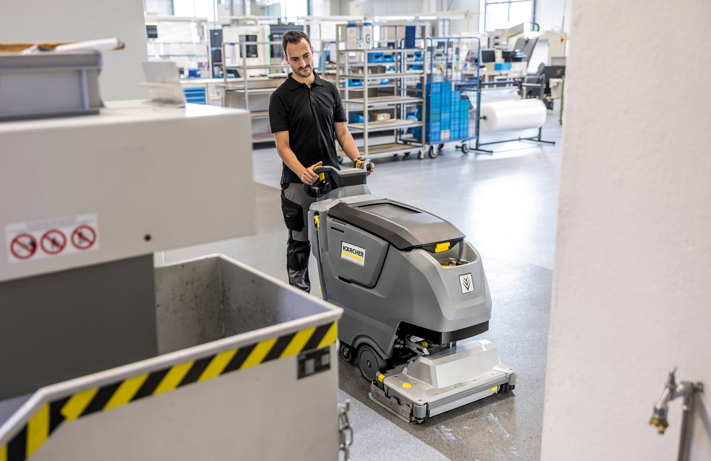 A worker uses a walk-behind scrubber to clean floors in a manufacturing plant.