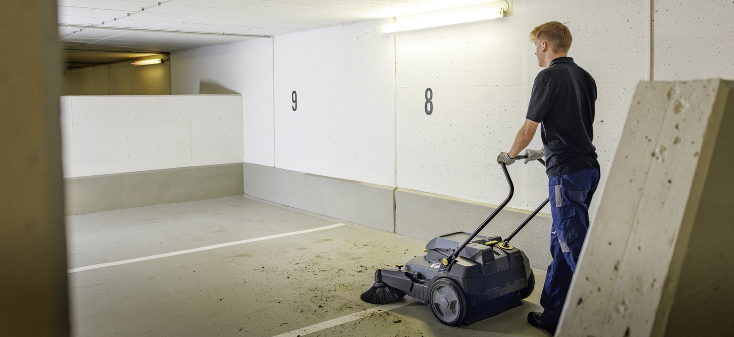 A person pushes a walk-behind sweeper through a parking garage