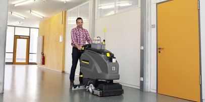 A worker uses a walk-behind floor scrubber in an office environment.