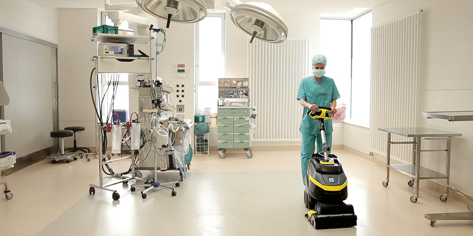 A worker in medical scrubs uses a compact walk-behind floor scrubber to clean operating room floors.
