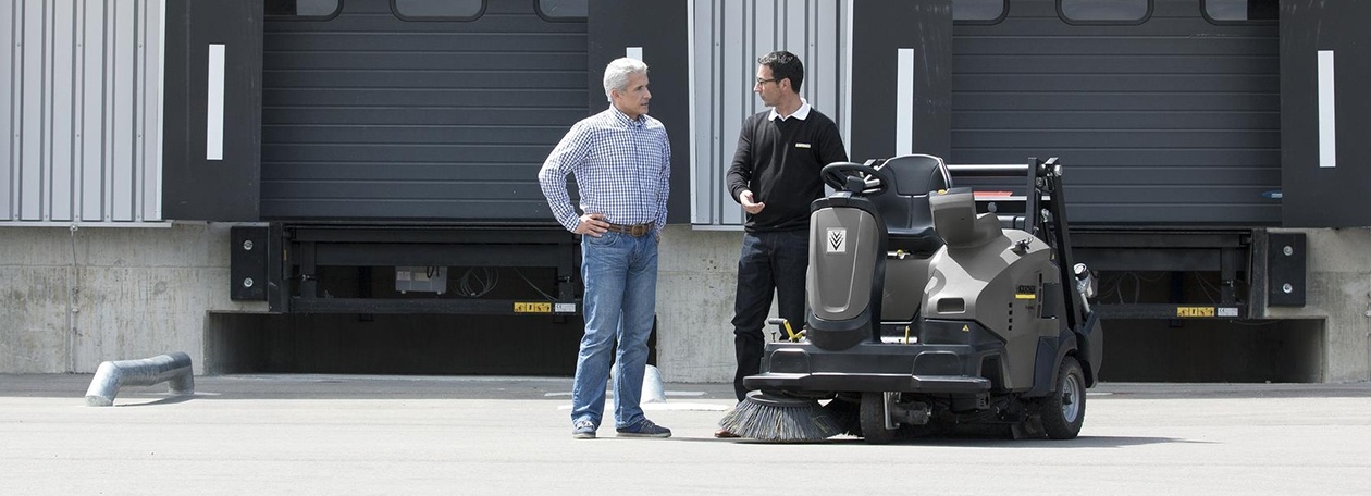 Certified Pre-Owned & Refurbished Equipment A Kärcher representative talks to a customer about a ride-on floor sweeper.