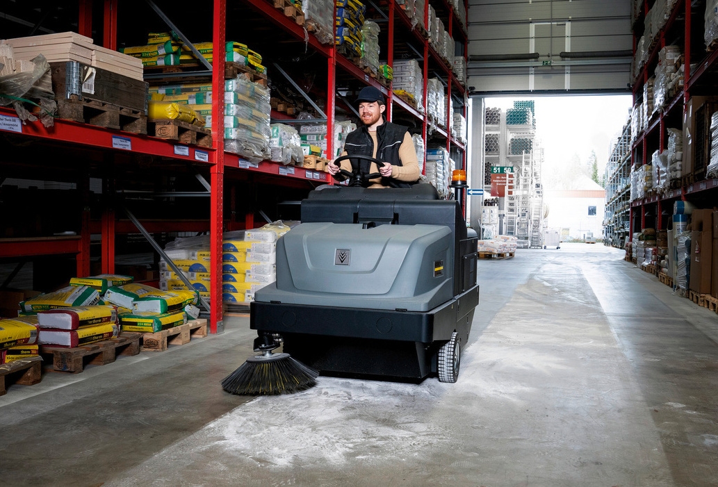 A man cleans a warehouse floor using a Kärcher ride-on floor sweeper