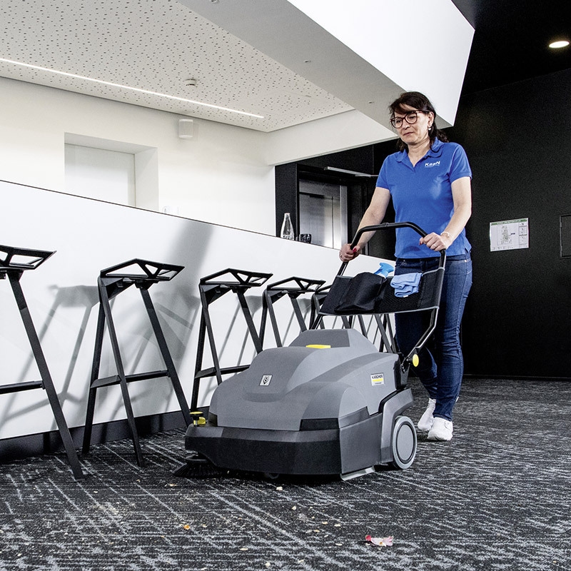 Productivity & Efficiency A worker sweeps carpet with the Kärcher KM 85/50 W walk-behind floor sweeper.