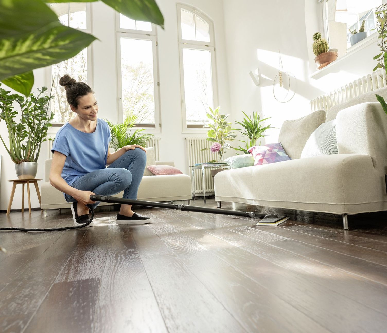 A woman clean cleans under a couch with a flat floor tool.