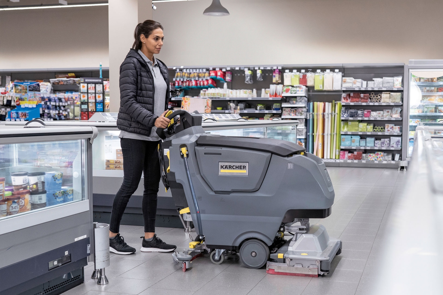 A worker cleans floors in a grocery store using a Kärcher walk-behind floor scrubber.