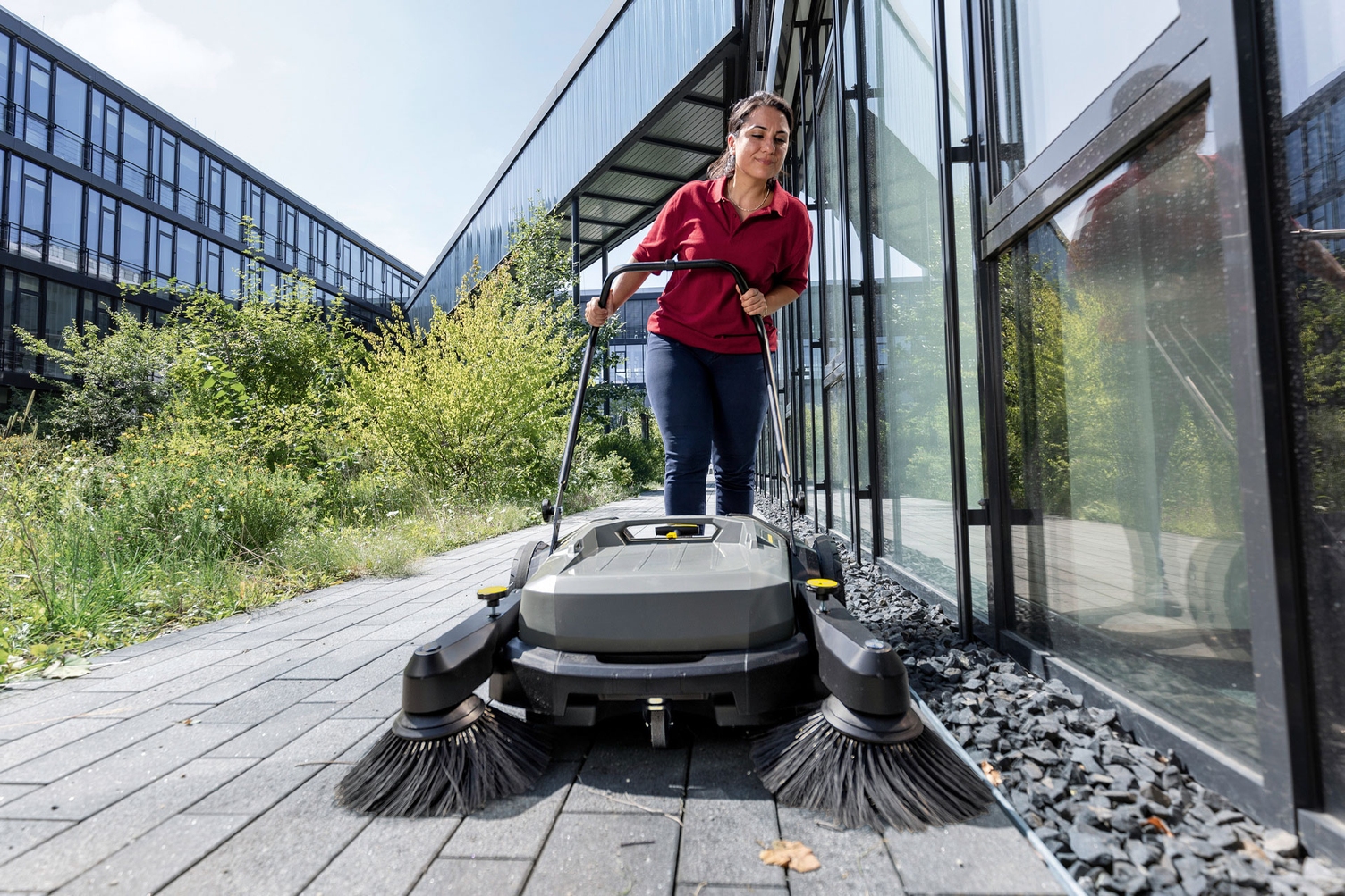 A worker uses a walk-behind floor sweeper to pick up debris outside a commercial building.