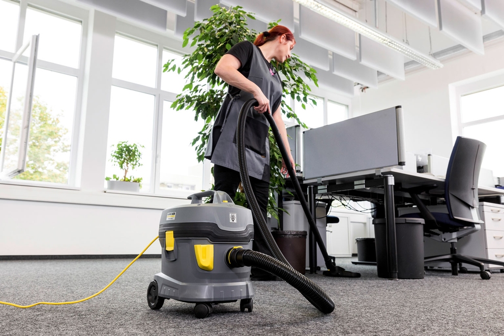 A worker cleans under desks using a canister vacuum.