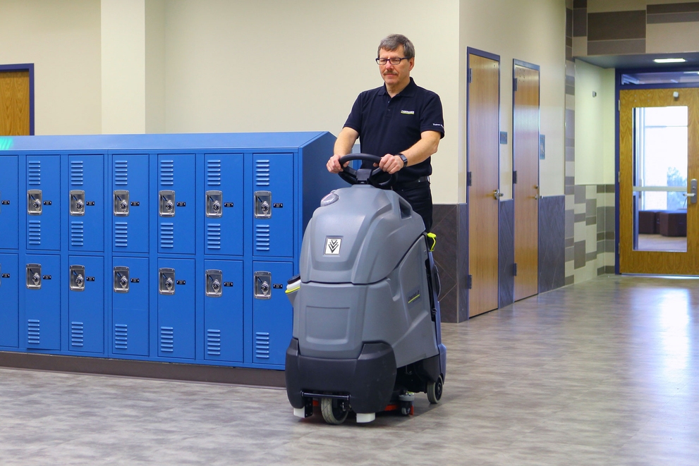 A worker cleans floors inside a school using a Kärcher Chariot™ stand-on floor scrubber.
