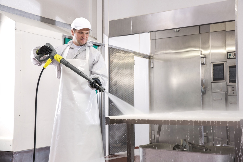 A worker cleans in an industrial kitchen with an electric-powered hot water pressure washer.