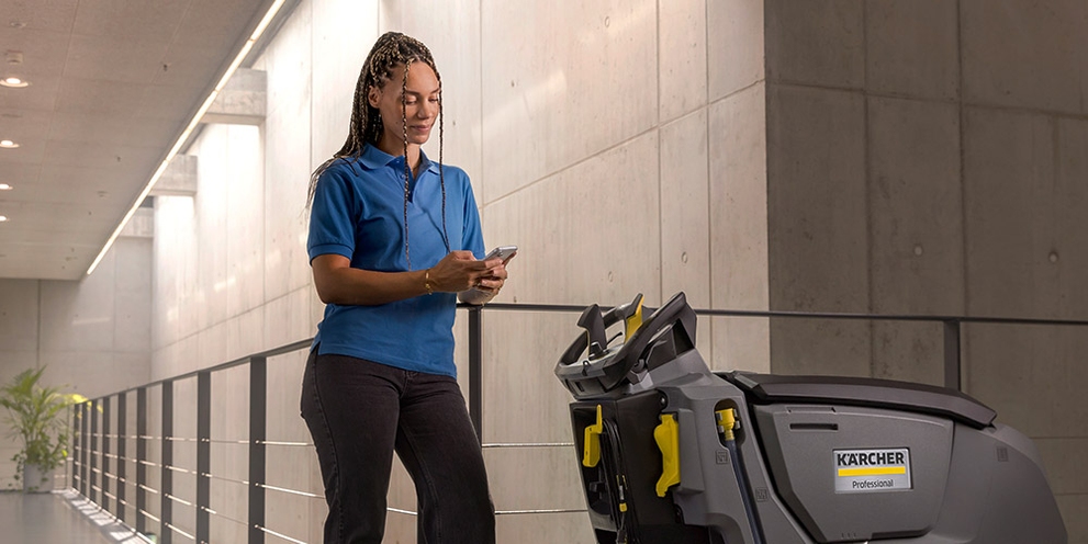 A woman reads from her smartphone while standing next to a walk-behind floor scrubber.
