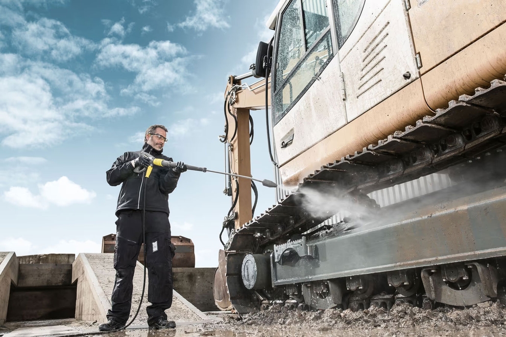 A worker cleans construction equipment with a gas-powered hot water pressure washer.