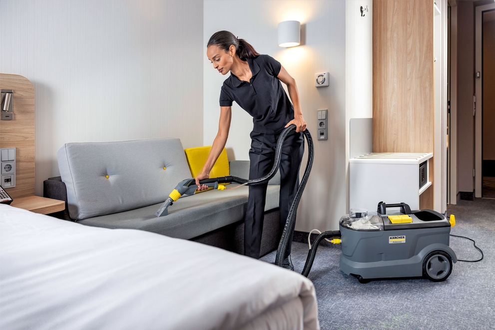 A worker cleans a couch in a hotel room using a compact extractor.