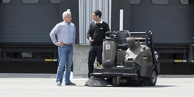 Two men talk beside an industrial ride-on floor sweeper.