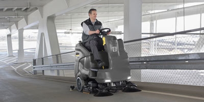 A worker uses a ride-on floor scrubber to clean a parking garage.
