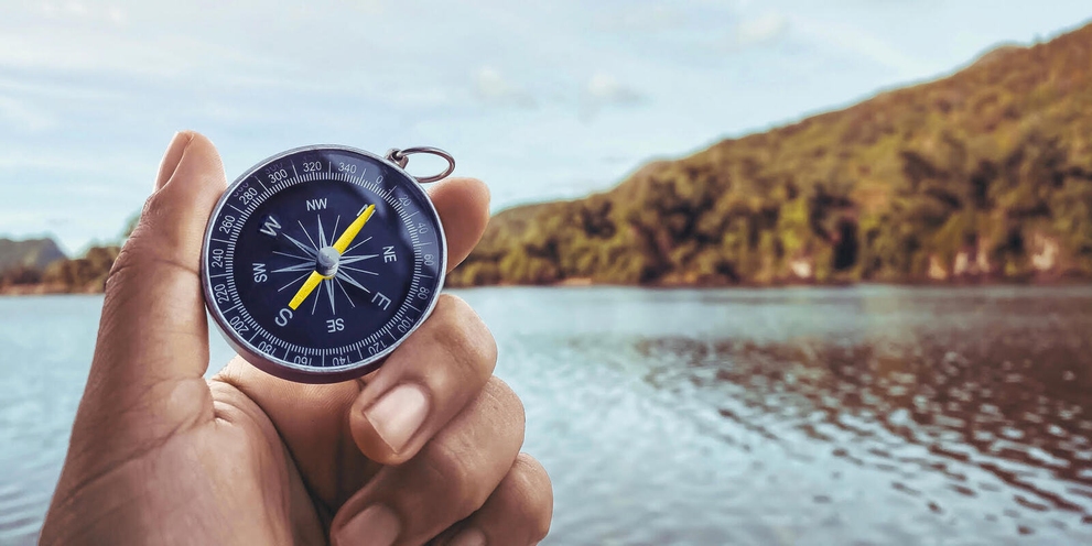A hand holds a compass with a mountain and body of water in the background