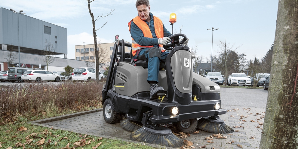 A worker uses a ride-on floor sweeper to clean outdoor areas.