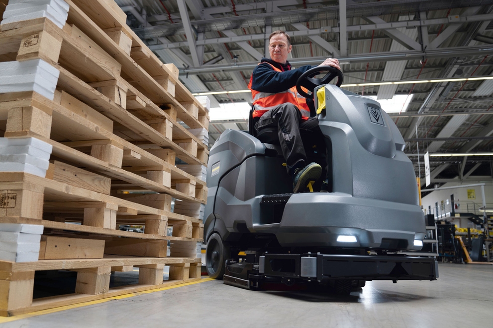A worker cleans warehouse floors with a Kärcher ride-on floor scrubber.
