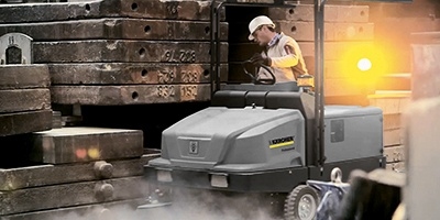 A worker uses a ride-on floor sweeper to clean inside a factory.