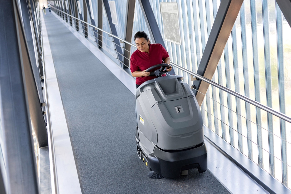 A worker cleans a long corridor using a stand-on vacuum.