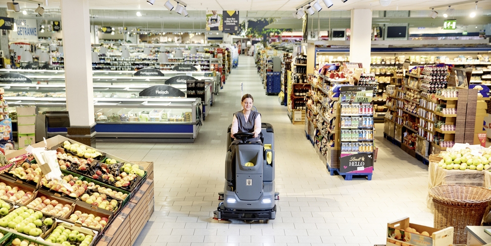 A worker uses a ride-on floor scrubber to clean grocery store floors.
